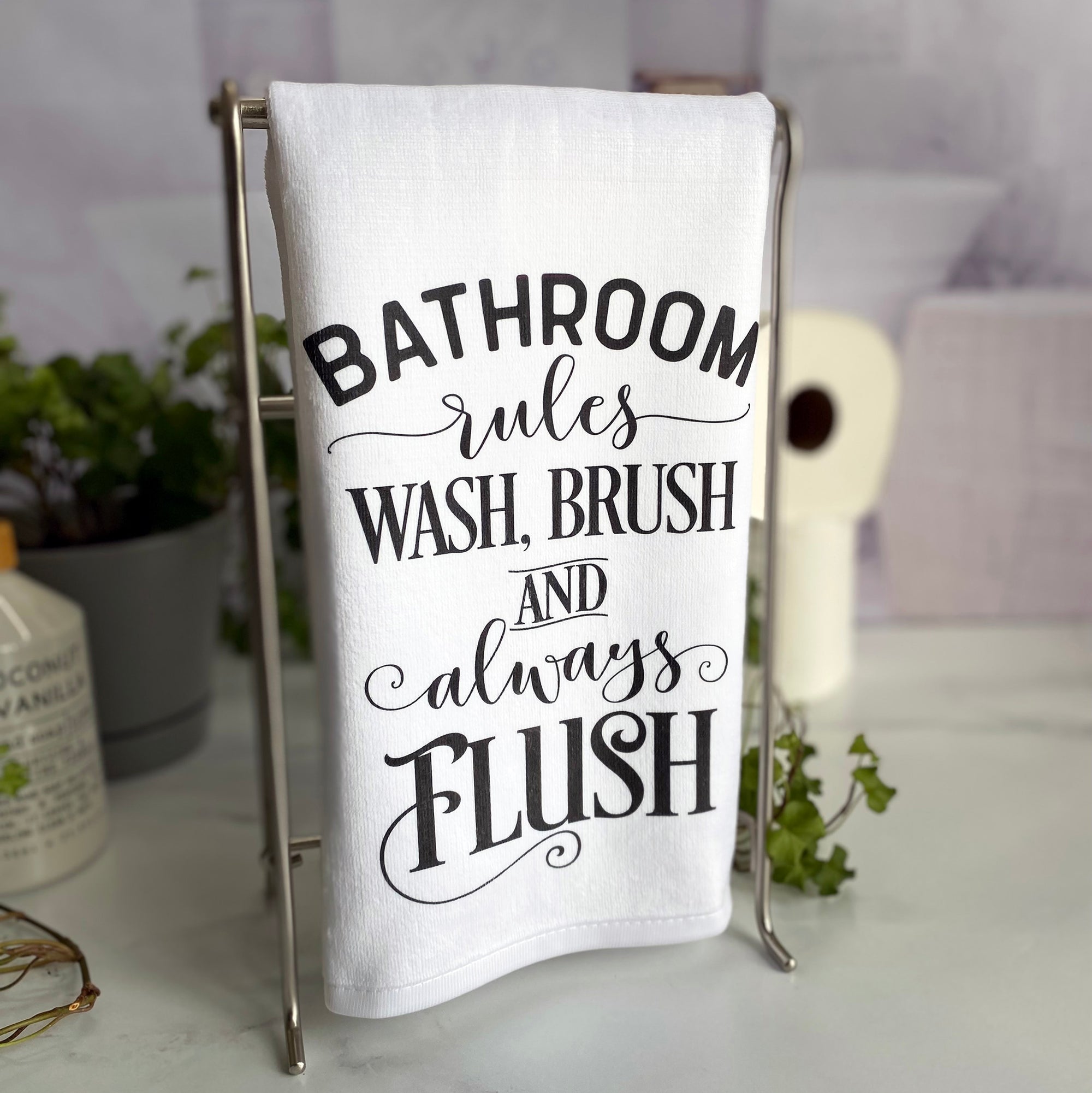 Bathroom Rules Hand Towel hanging in front of a houseplant, a stack of toilet paper rolls, and a bottle of hand soap