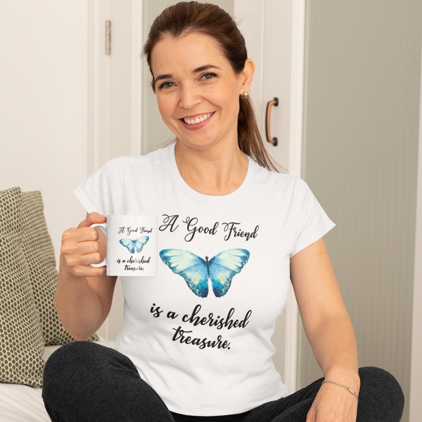Smiling woman wearing a white Tshirt with a beautiful blue watercolor butterfly and the words, "A Good Friend is a cherished treasure.", holding a matching ceramic mug.