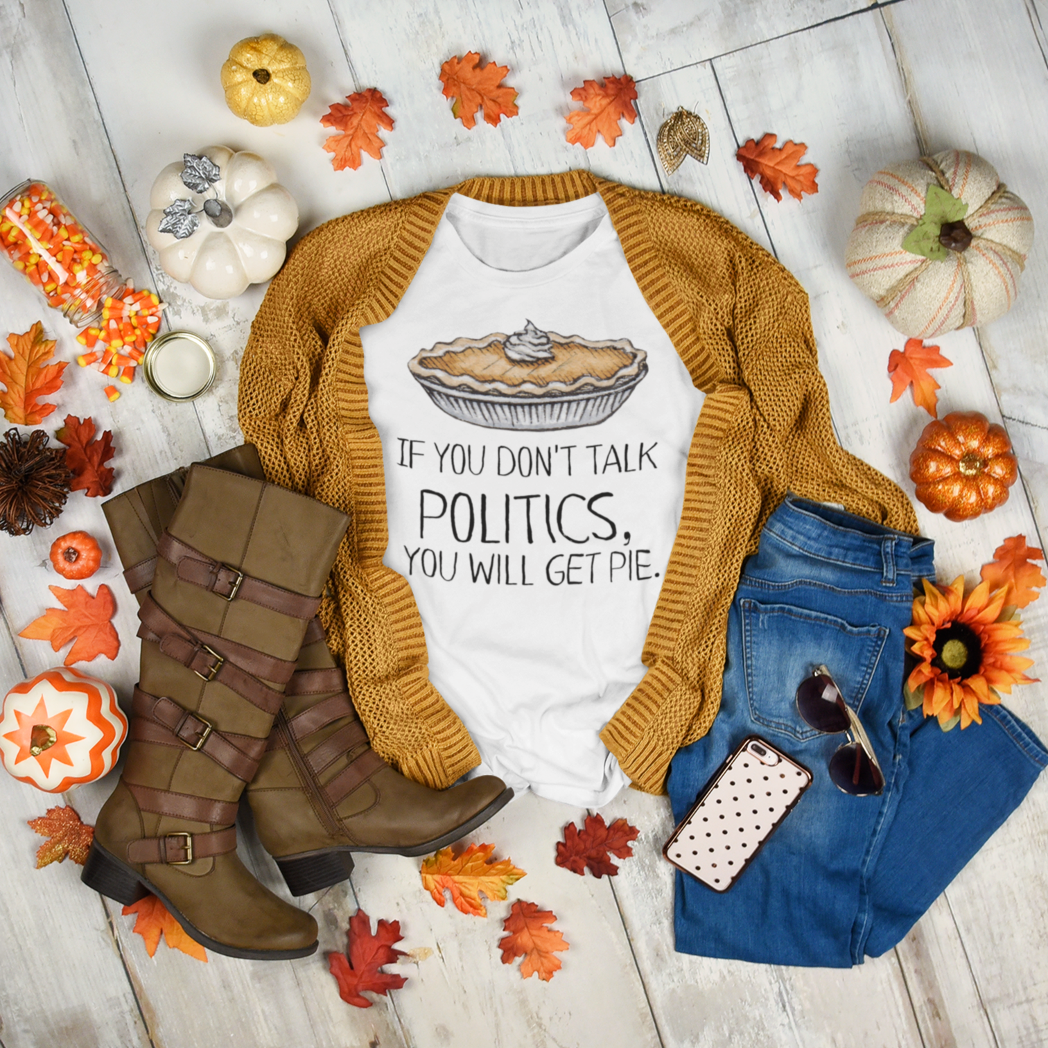 Beautiful flat lay display with a white 'If You Don't Talk Politics, You Will Get Pie' tee with a watercolor pumpkin on the chest and a pumpkin colored cardigan sweater, tall brown boots, blue jeans, and an array of fall leaves, pumpkins and candy corn.