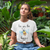 Young woman wearing a white "You are the Bee's Knees" tee with a cute watercolor bee in place of the word 'bees'.  She's sitting on the floor surrounded by plants.