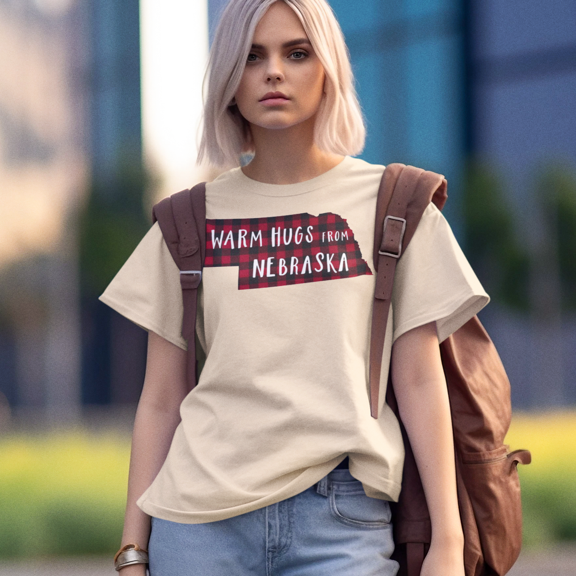 Young woman wearing a beige oversized T-shirt with a red and black buffalo plaid design in shape of Nebraska with the words, "Warm Hugs from Nebraska" on the chest.  She's wearing an oversized brown leather backpack and light denim jeans and a silver bracelet.