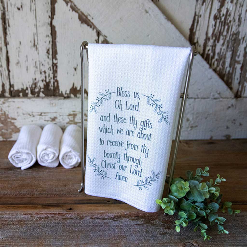 Beautiful waffle dishtowel with the Catholic prayer, "Bless Us, Oh Lord and these thy gifts which we are about to receive from thy bounty through Christ our Lord. Amen" This towel is hanging in front of a barn door with several other towels rolled up in the background and a succulent sprig in the foreground.