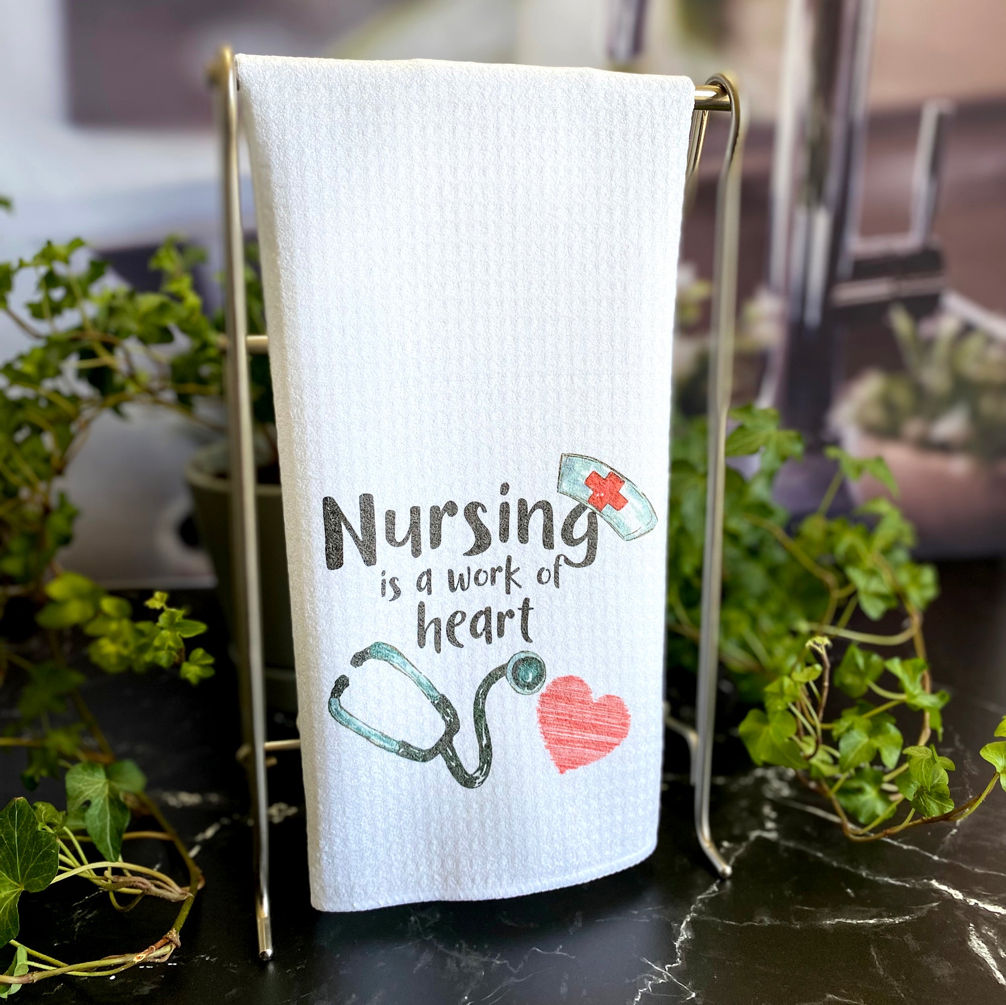 Dishtowel with "Nursing is a work of heart" with image of a hand drawn stethascope, heart and an old timey nurse's cap that appears to be hanging on the letter G.  
