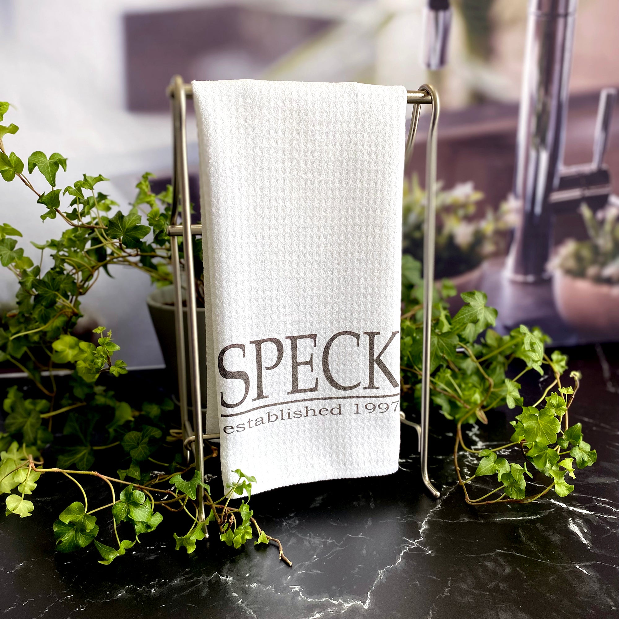 A White waffle dishtowel with a last name in all capital letters in a serif font with a line under the name and the established year below that.  The towel is hanging form a metal stand, surrounded by ivy on a black marble surface.