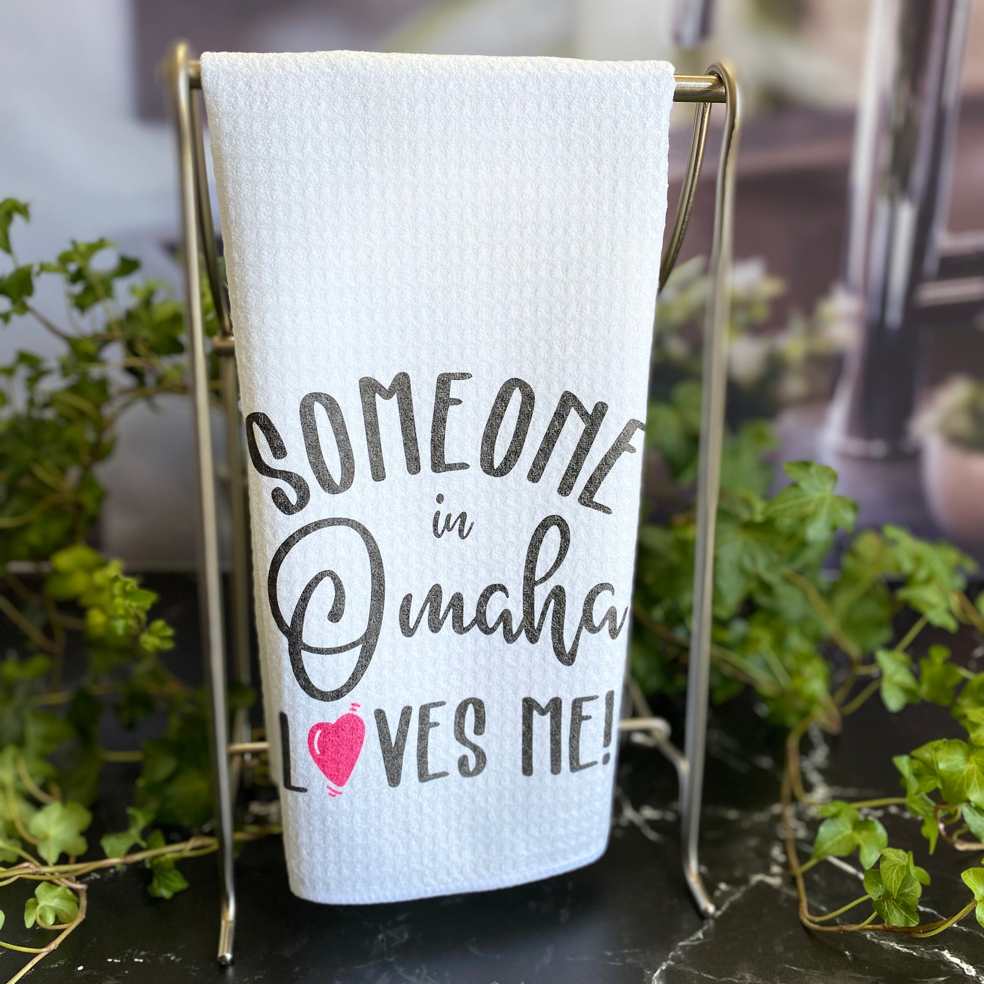 Someone in Omaha Loves Me! Dishtowel hanging on a silver stand surrounded by ivy.