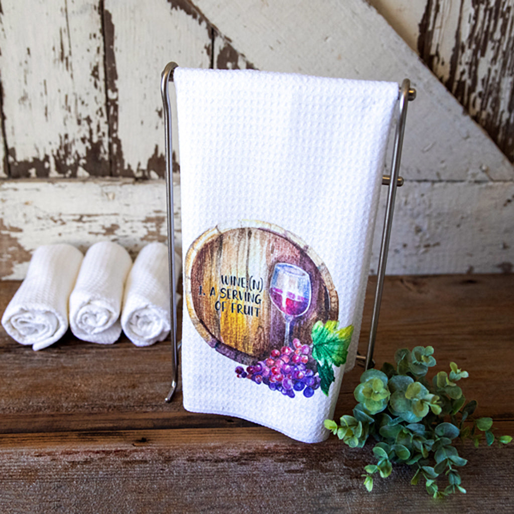 Dishtowel hanging in front of a rustic barn door.  The towel has a watercolor wine barrel, grapes and a glass of red wine and the words, "Wine(N) 1. A Serving of Fruit."