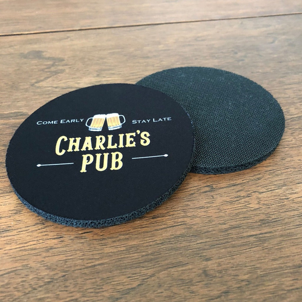 A pair of black fabric coasters with beer mugs that say, "Charlie's Pub- Come Early- Stay Late"