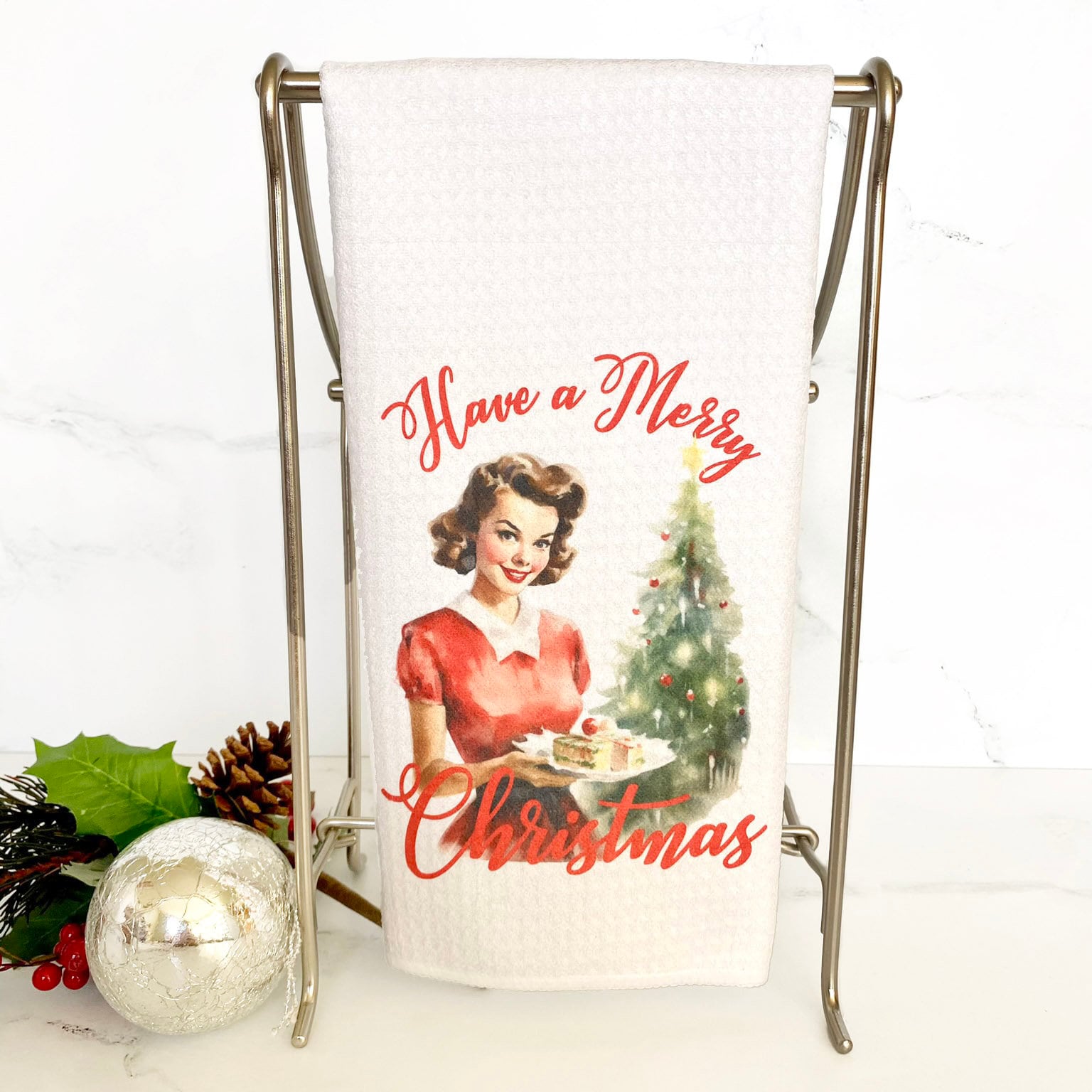 Have a Merry Christmas dishtowel with a red cursive font and a watercolor image of a woman holding baked goods with a Christmas Tree in the background.