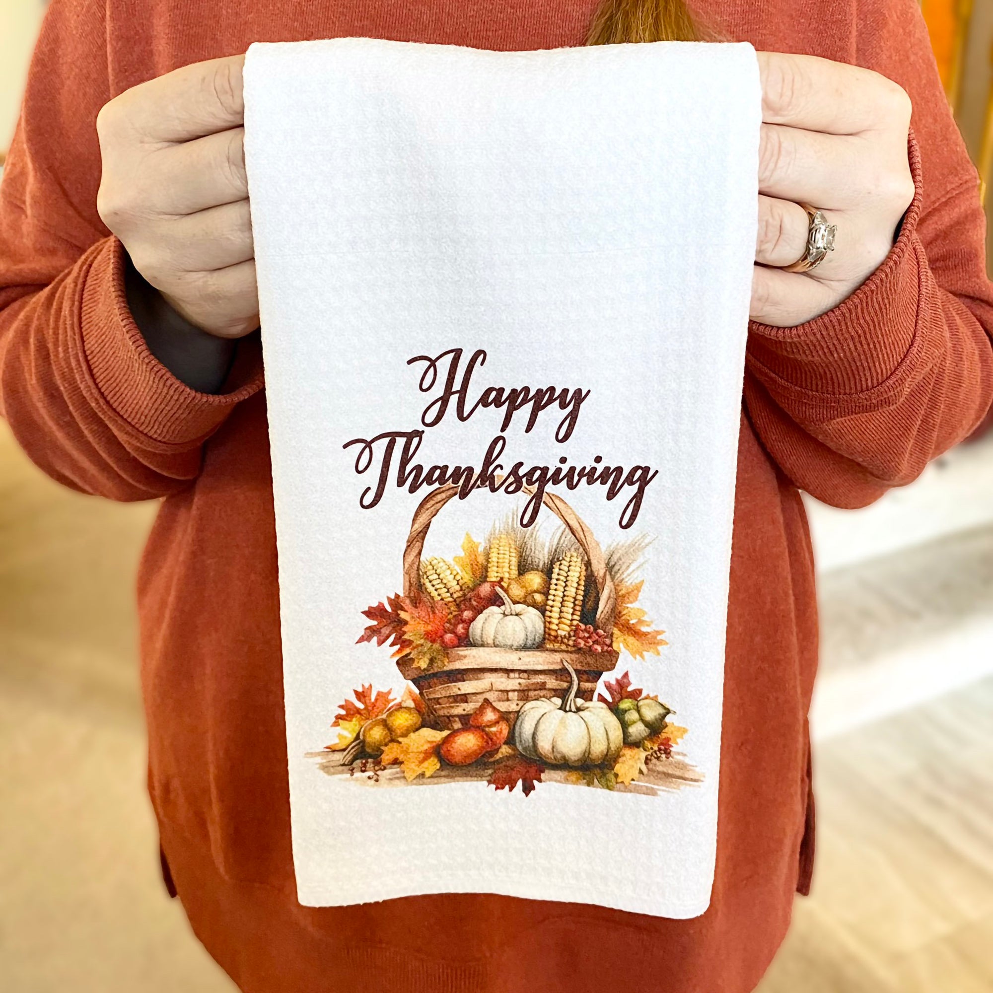 Woman in an orange sweater holding a white dishtowel adorned with a basket full of corn, fall leaves, pumpkins and gourds and the words, &quot;Happy Thanksgiving&quot; in a dark brown cursive font.