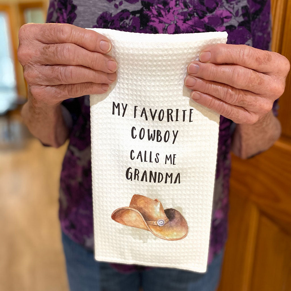 Elderly woman's hands holding up a dishtowel that says, "My Favorite Cowboy Calls Me Grandma" with a watercolor cowboy hat printed on it.